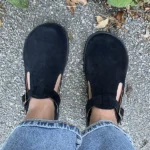 Black barefoot Mary Jane shoes with ankle straps, worn with jeans on an outdoor pavement.