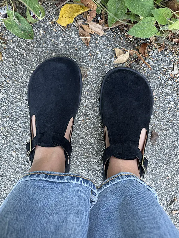 Black barefoot Mary Jane shoes with ankle straps, worn with jeans on an outdoor pavement.