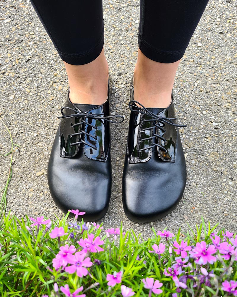 Black barefoot Oxford-style shoes with wide toe box, worn with black leggings, standing on a path next to bright pink flowers.