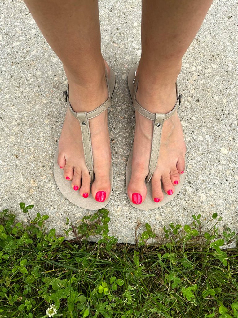 Barefoot sandals with thin metallic straps around the toes and ankles, worn on feet with red toenail polish, standing on a concrete path next to green grass.