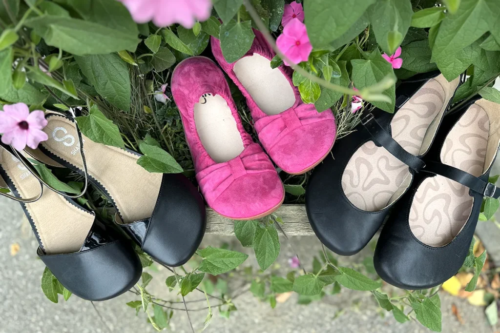 Three pairs of barefoot ballet flats in black and pink displayed on a wooden bench surrounded by greenery and flowers.