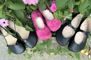 Three pairs of barefoot ballet flats in black and pink displayed on a wooden bench surrounded by greenery and flowers.