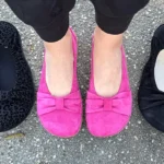 Feet wearing bright pink Bär Shoes Elly ballet flats surrounded by two pairs of black flats on pavement.