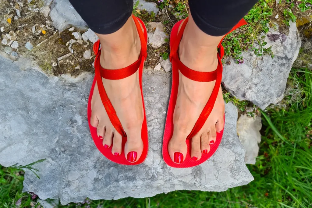 Top view of bright red Panta barefoot sandals with toe loop, worn on feet standing on a rock surrounded by grass