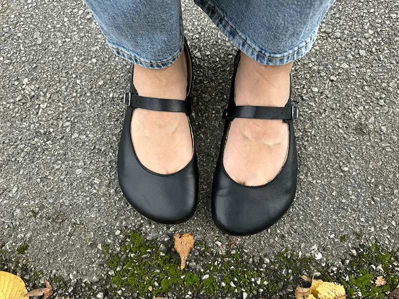 Top view of feet wearing black Jenon Villet barefoot Mary Jane flats on pavement.