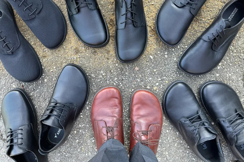 Top-down view of multiple barefoot men’s dress shoes arranged in a circle on asphalt, with one person wearing brown shoes in the center.