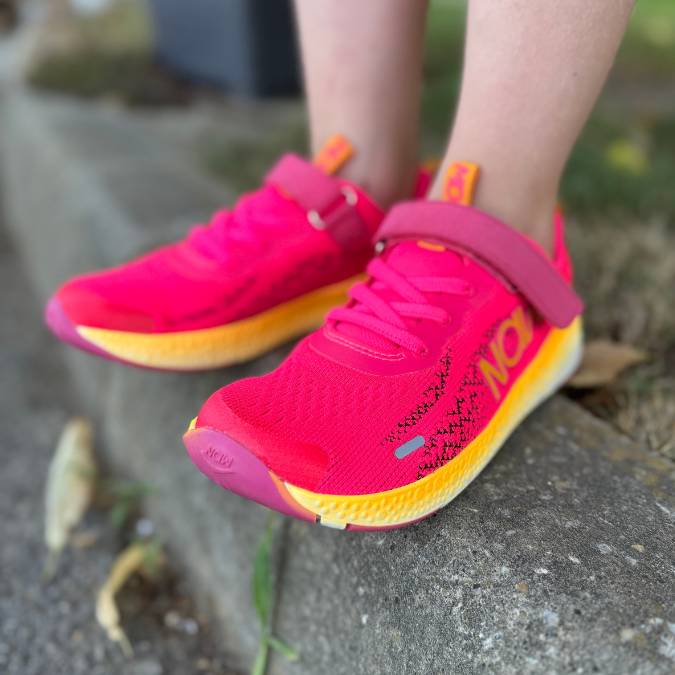 Bright pink and yellow NAW barefoot shoes with mesh uppers and a Velcro strap, worn by a child standing on a curb.
