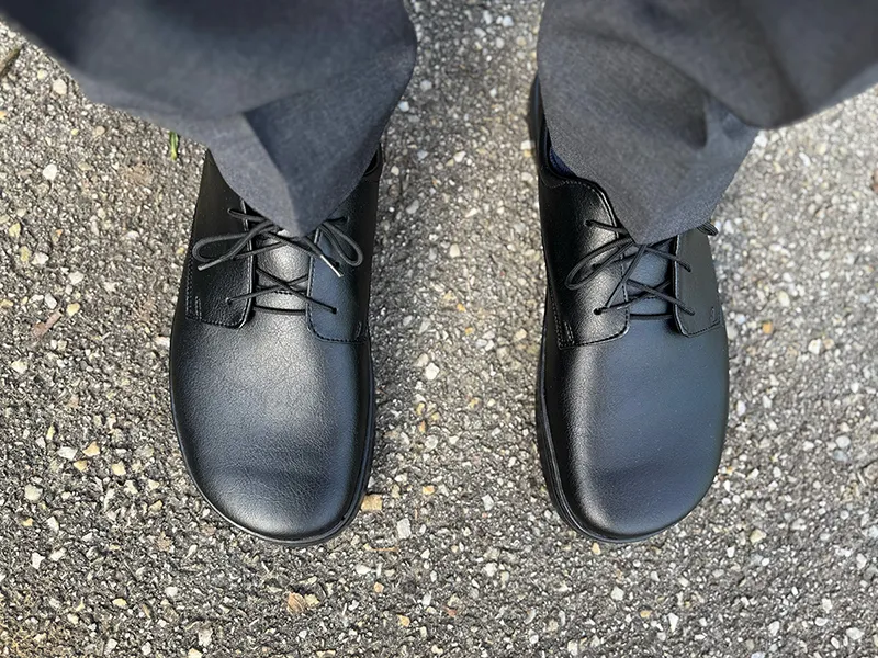 Peerko barefoot dress shoes in black leather, paired with formal grey trousers, photographed from above to show the wide, foot-shaped toe box and minimal sole.
