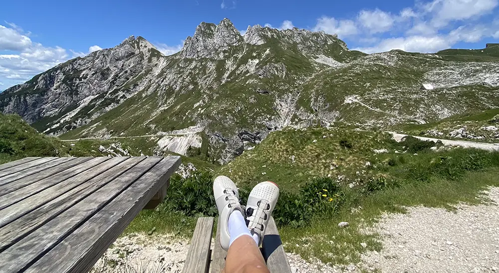 Cyclist resting with feet up on a bench, wearing Strong Feet Athletics wide toe box cycling shoes, overlooking a scenic mountain landscape under a bright blue sky.