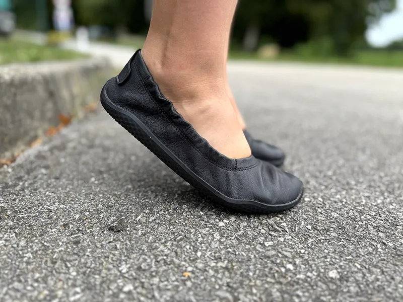 Side view of feet wearing black VIVOBAREFOOT ballet flats on asphalt, showing flexible sole during heel lift.