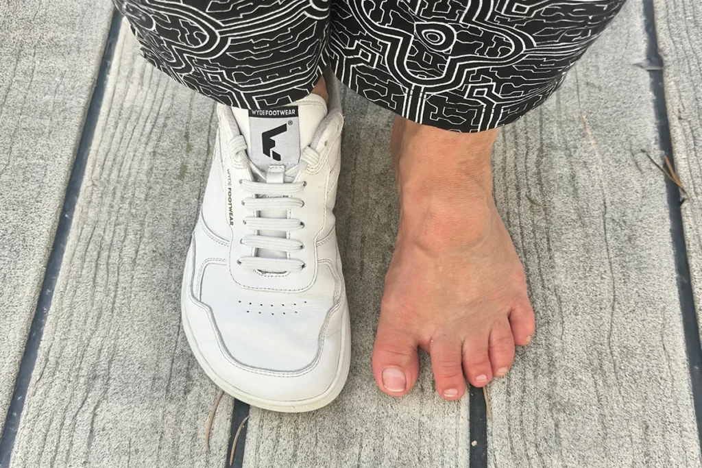 Bare foot next to a Wyde Footwear Nova sneaker on wooden planks, highlighting the shoe’s natural foot shape and wide toe box.