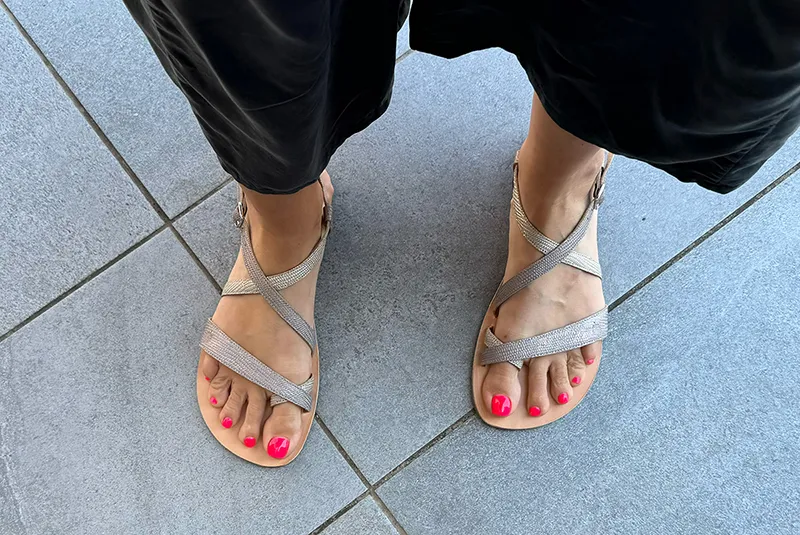 Close-up of feet wearing Lang.S Selene barefoot sandals in metallic silver with criss-cross straps and ankle buckles, shown on a tiled floor
