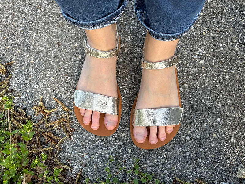 Metallic gold Saguaro Glow I barefoot sandals with wide toe box and ankle strap, worn on bare feet, standing on textured pavement.