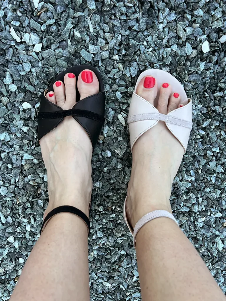 Close-up of feet wearing Shapen Petal barefoot sandals in black and blush pink, with open toe design and ankle straps, shown on a gravel background.