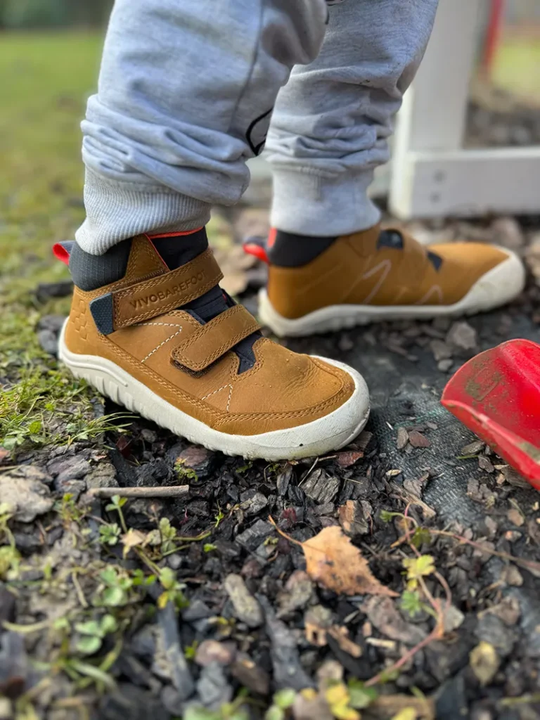Child wearing brown Vivobarefoot barefoot sneakers with Velcro straps outdoors on grass and wood chips
