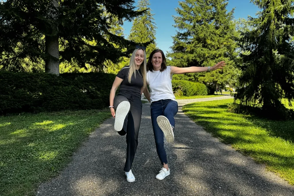 Two smiling women standing on a sunny park path, balancing playfully on one leg and showing the soles of their white barefoot sneakers.