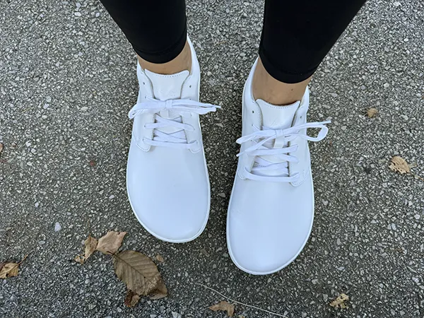 White barefoot sneakers with wide toe box, worn with black leggings on concrete ground.