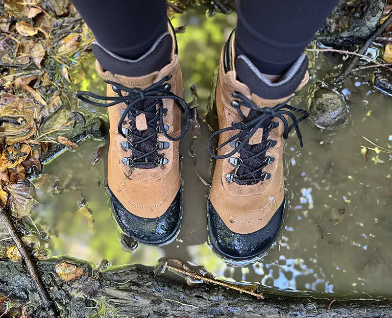 Brown and black waterproof barefoot hiking boots standing in a muddy puddle on a forest trail.