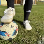 White barefoot soccer cleats stepping on the ball near the sideline.